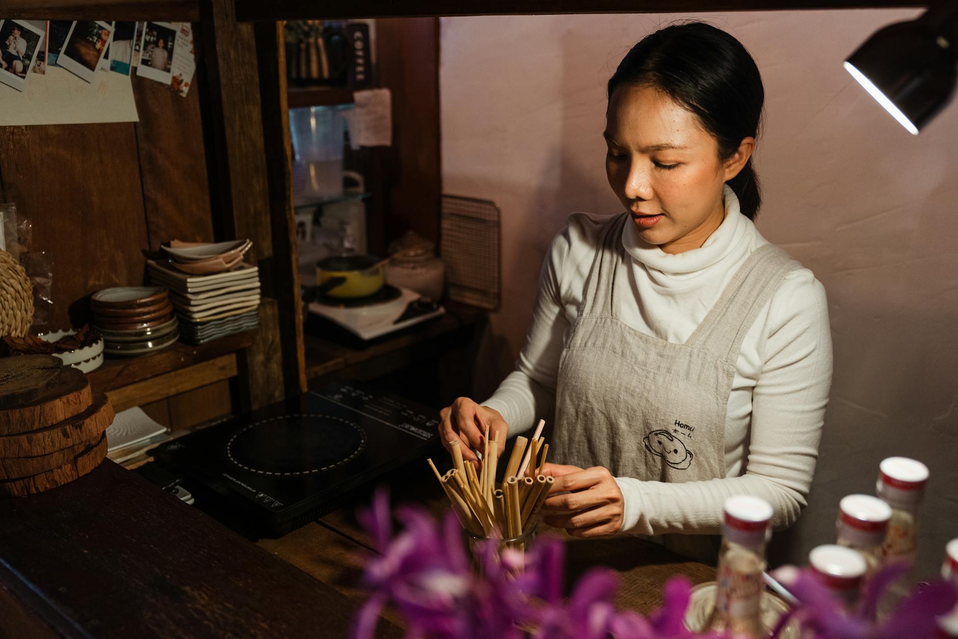 Frome above of busy young ethnic female bartender wearing apron standing behind counter with alcohol bottles and adjusting wooden straws for cocktails while looking down in cafe with rustic wooden interior in dark light in evening